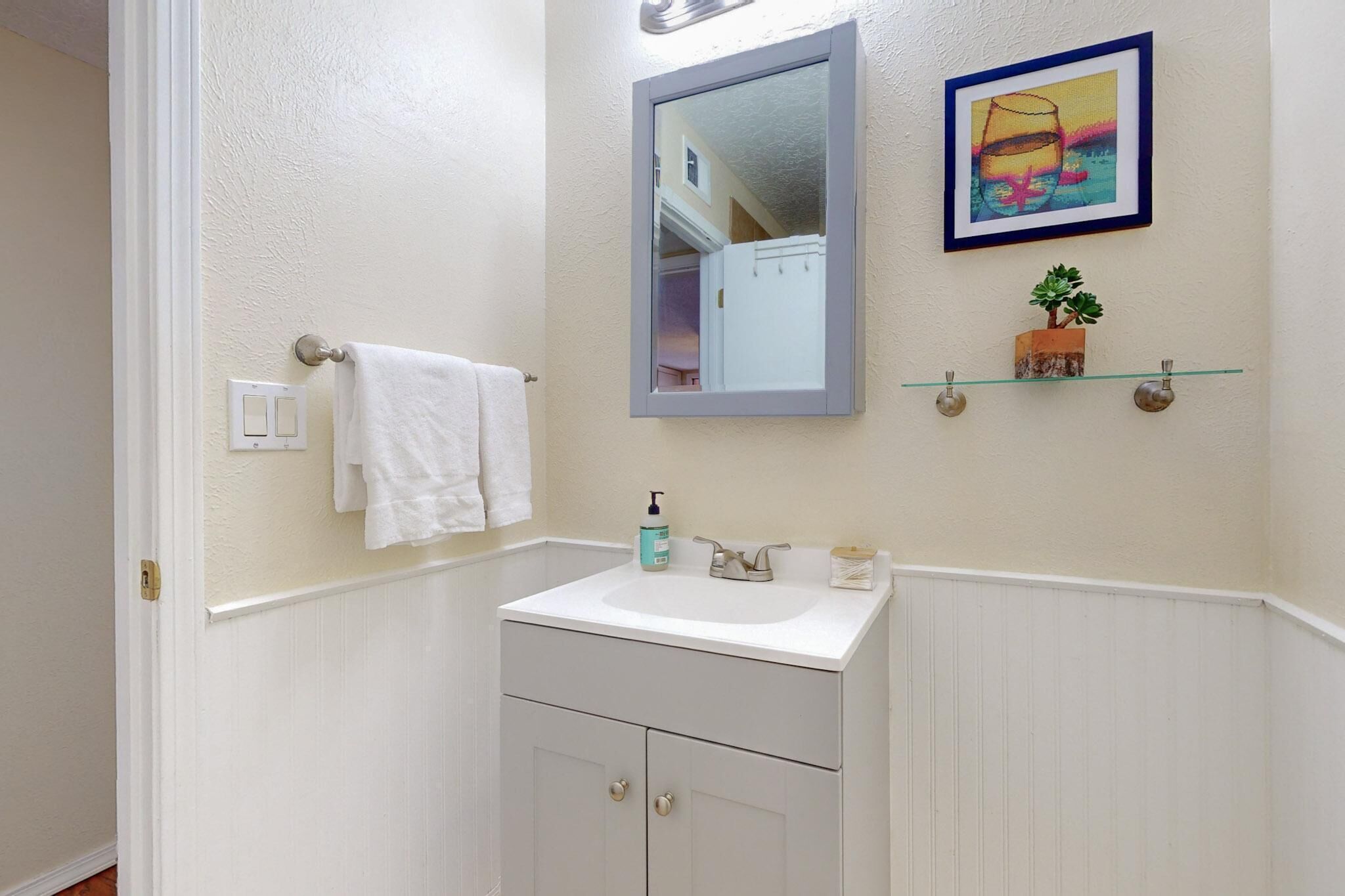 Une salle de bain avec un meuble-lavabo blanc, un miroir, un porte-serviettes, une étagère avec une plante et un tableau encadré au mur.