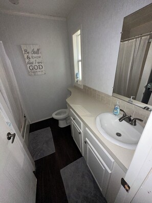 Une petite salle de bain avec des toilettes, un lavabo et un miroir. Un panneau est accroché au mur, sur lequel on peut lire : « Soyez calme et sachez que je suis DIEU. »