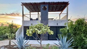 Une maison moderne à deux étages, dotée d’un patio couvert, d’un pilier en béton et d’un jardin agrémenté de rochers et de plantes.