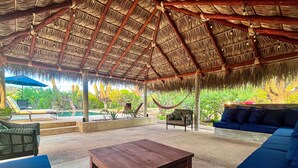 Un espace extérieur aménagé, abrité et doté d’un toit de chaume, avec des fauteuils confortables et une piscine en arrière-plan.