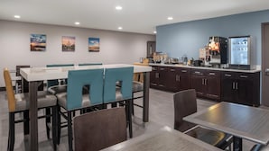 A hotel dining area with a long table, blue cushioned chairs, and a kitchen area with a refrigerator and coffee machine.