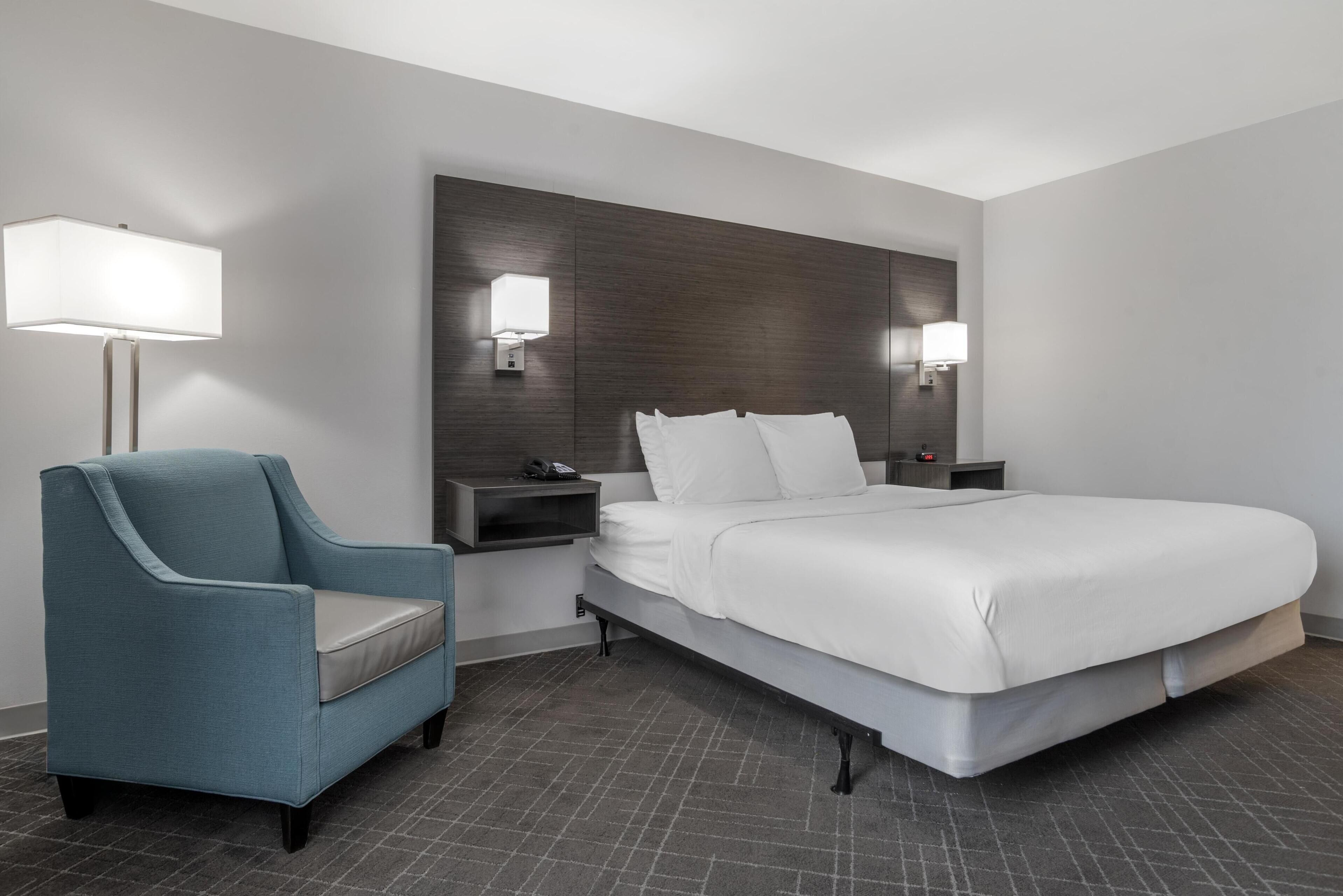 A modern hotel room with a large bed, a blue armchair, bedside tables, and wall-mounted lamps.