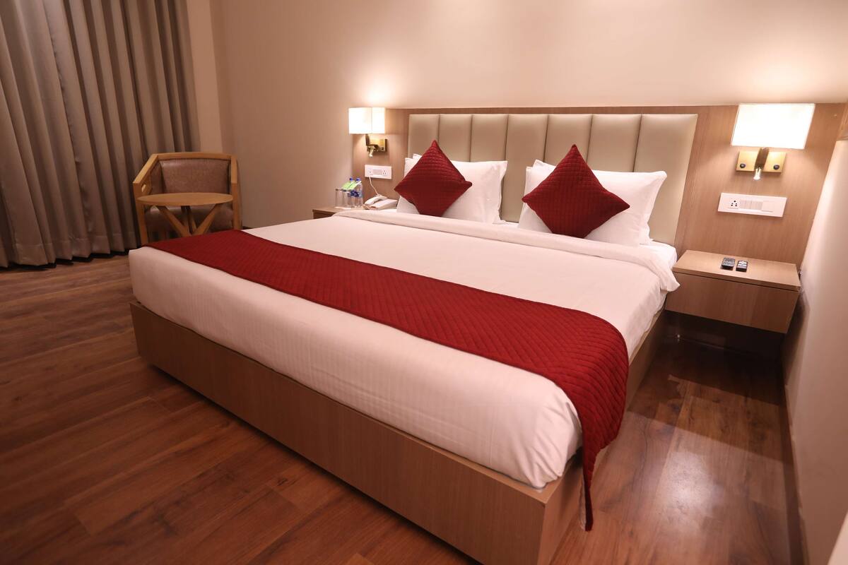 Una habitación de hotel con una cama grande, suelo de madera, una silla, mesitas de noche con lámparas y una ventana con persianas.