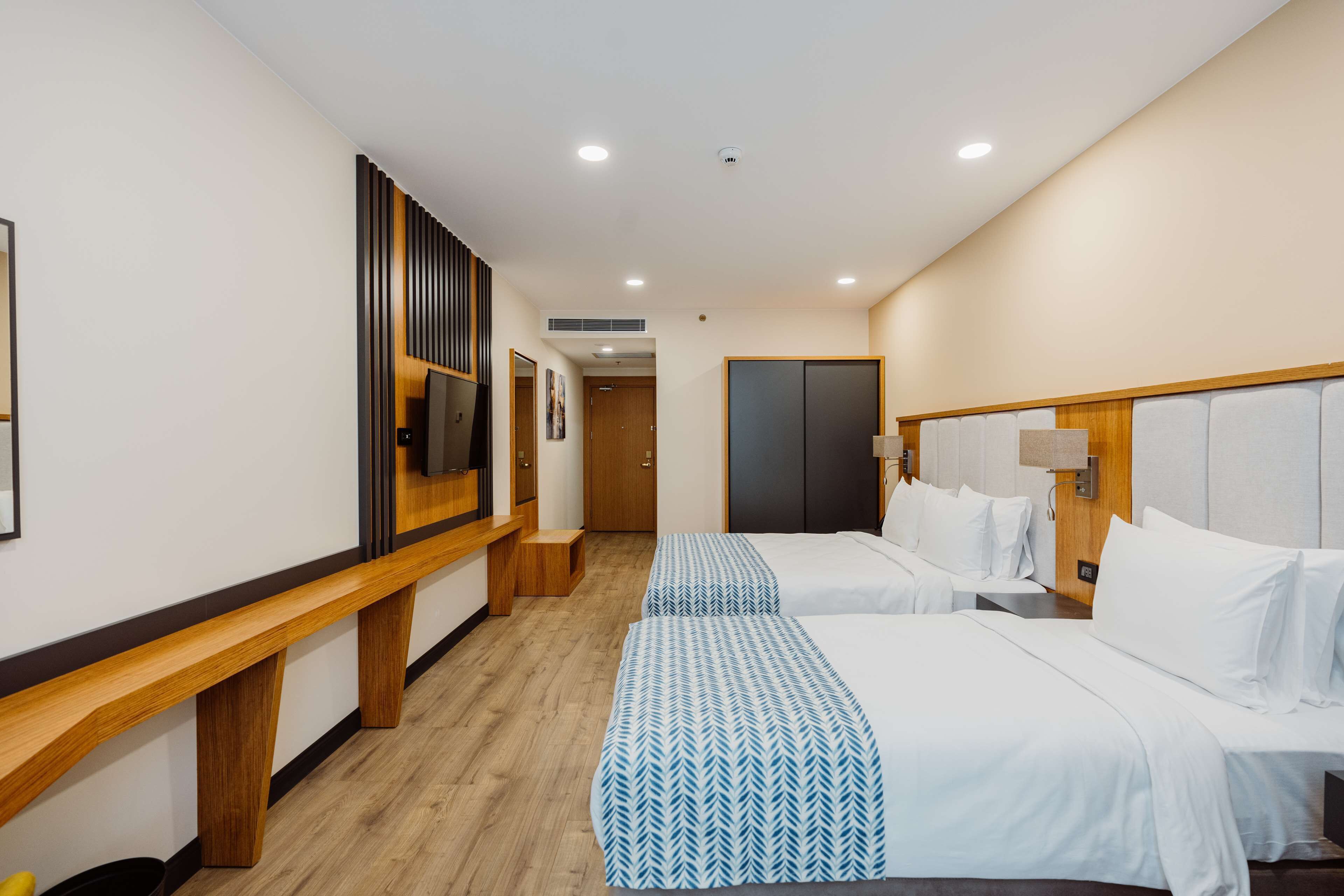 Habitación de hotel con dos camas, un banco de madera, un televisor y una puerta que da a otra habitación.