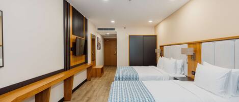 Habitación de hotel con dos camas, un banco de madera, un televisor y una puerta que da a otra habitación.