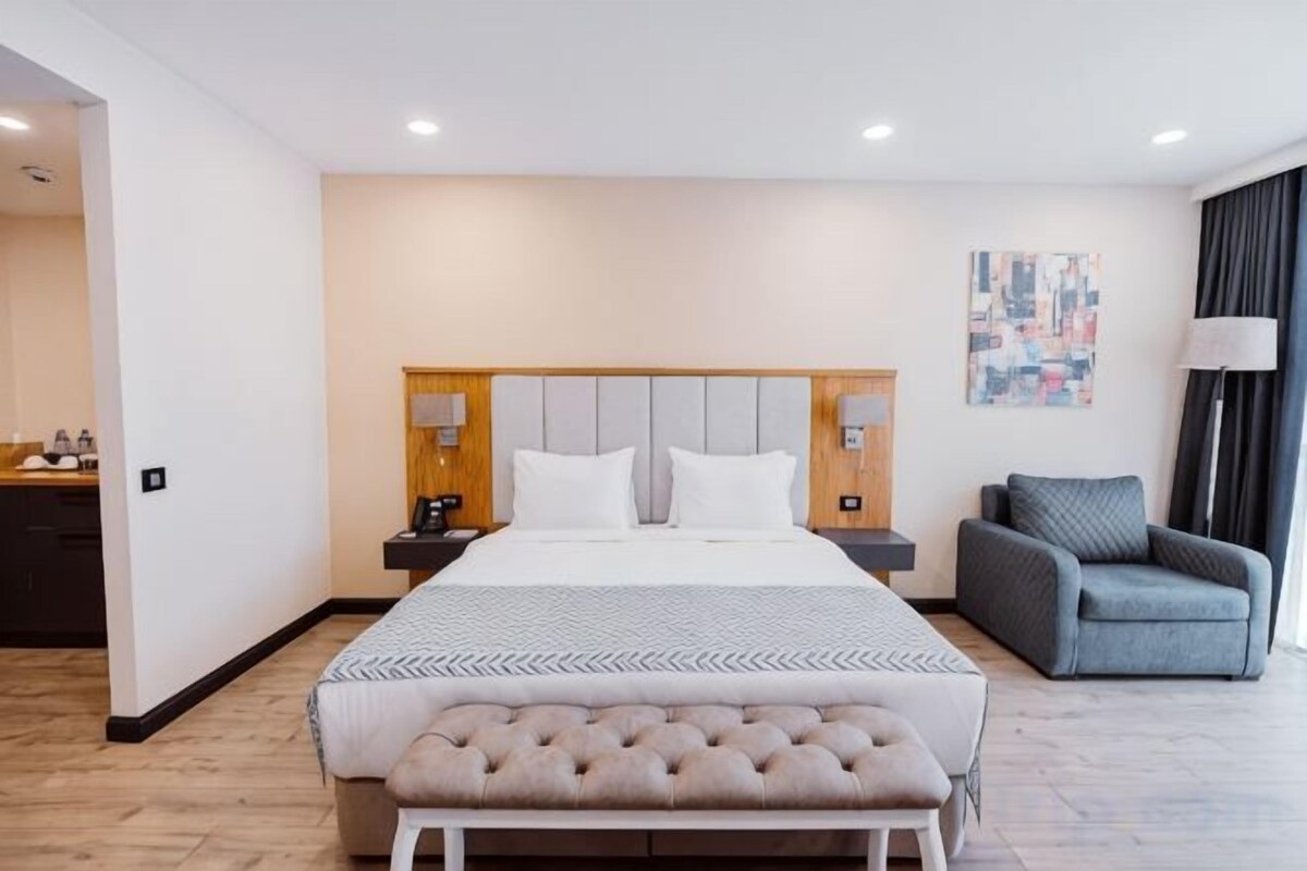 Una habitación de hotel con una cama grande, mesitas de noche, una silla y un banco.
