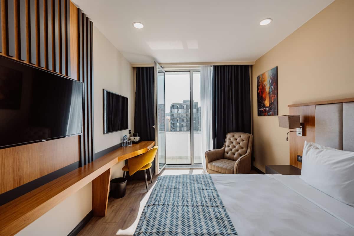 A hotel room with a bed, a desk, a chair, a TV, and a window with a view of buildings.