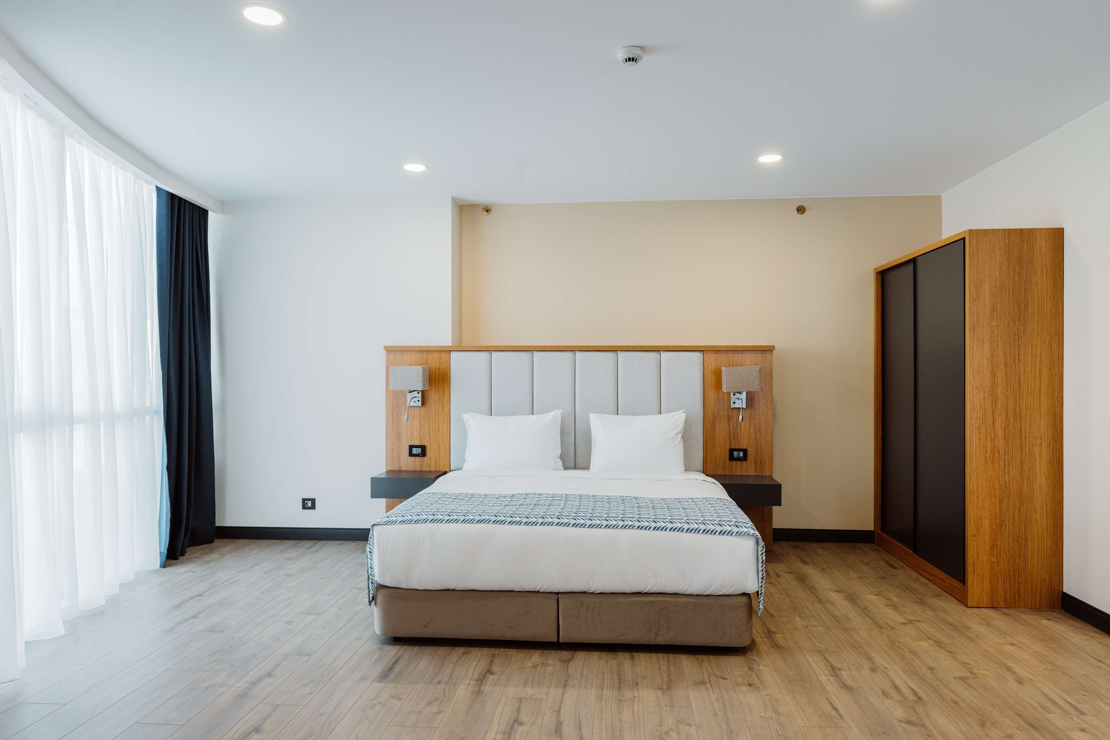 A modern hotel room with a large bed, wooden headboard, and a wardrobe.