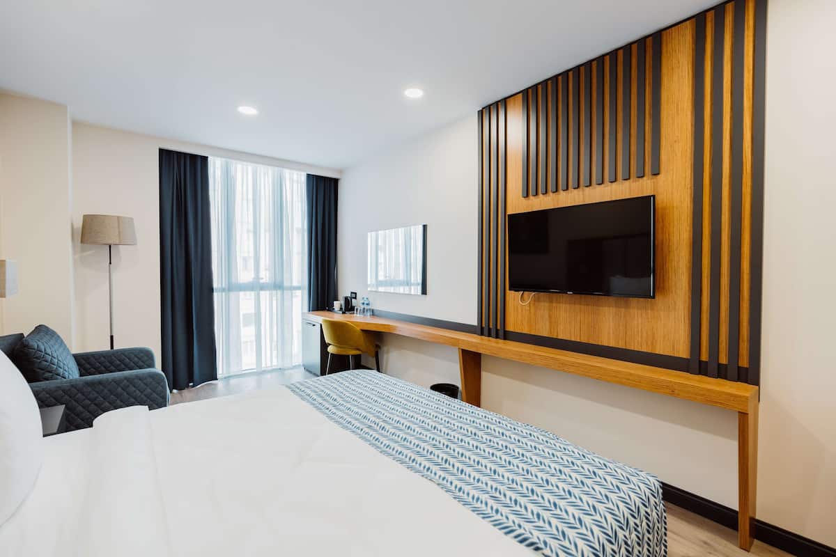 A modern hotel room with a large bed, a desk, a flat-screen TV mounted on a wooden panel, and a view of the city through large windows.