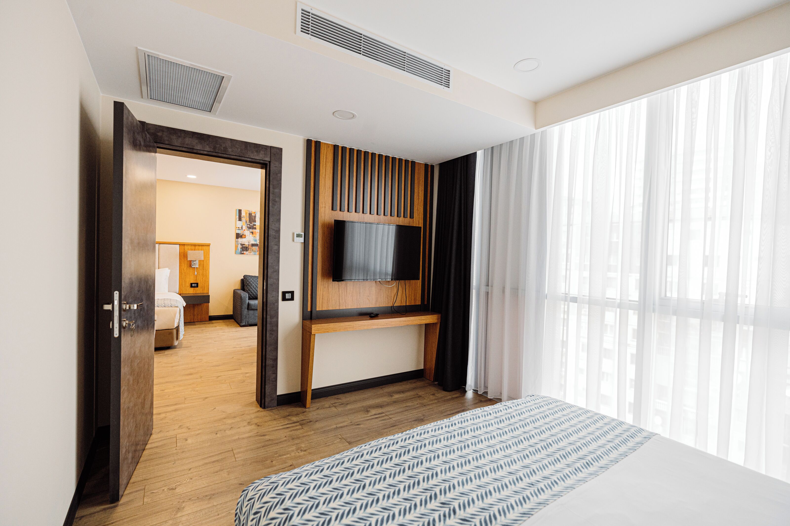 A hotel room with a bed, a TV mounted on a wooden panel wall, a large window with sheer curtains, and a door leading to another room.