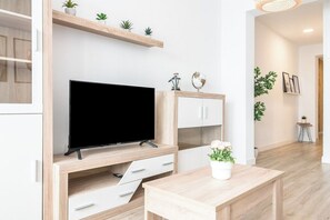 Un salon moderne avec un téléviseur à écran plat, des étagères en bois ornées de plantes, un meuble blanc et une petite table sur laquelle est posée une plante en pot.