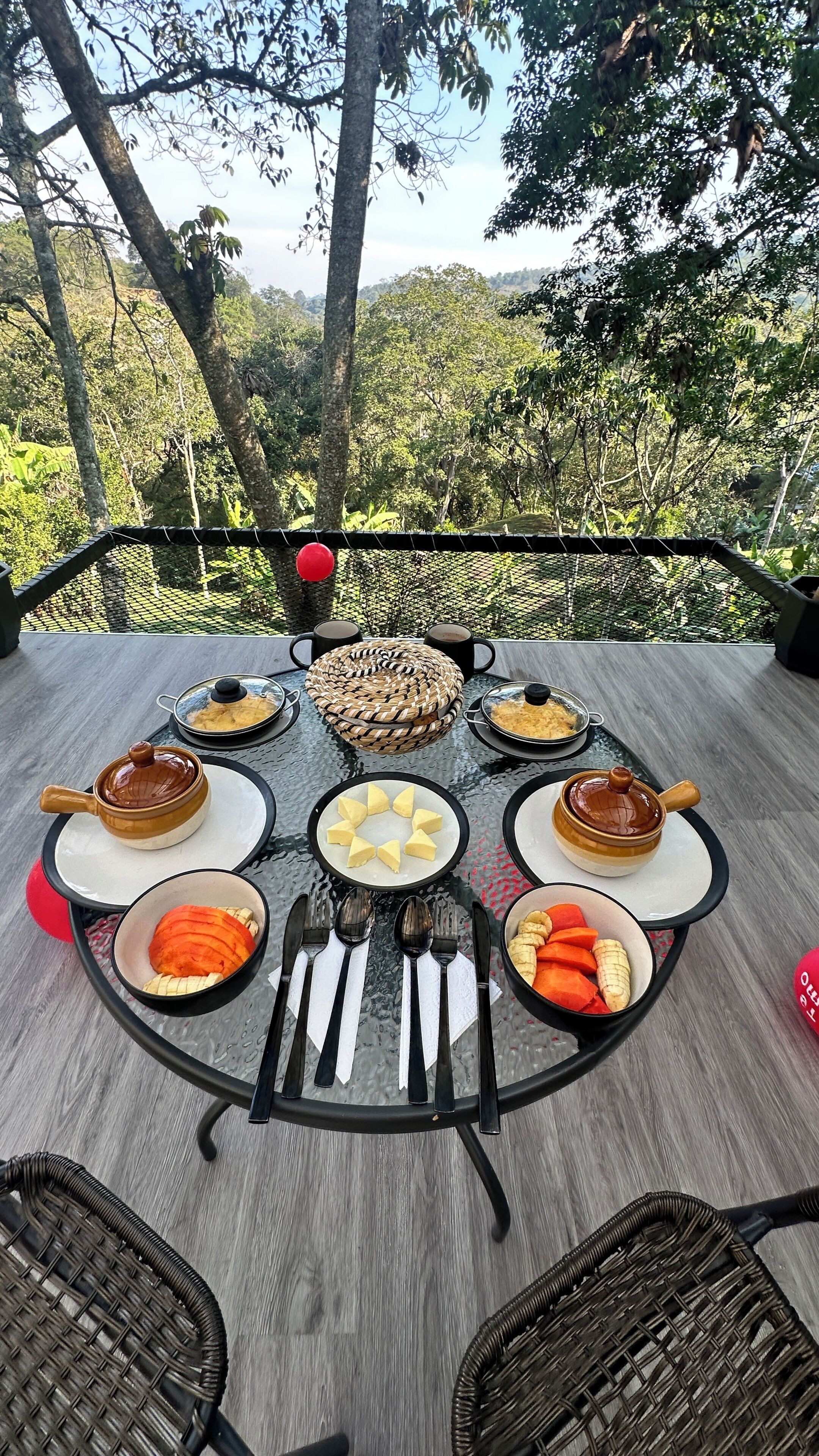 A round table with a glass top, surrounded by chairs, set outdoors with a view of trees and hills.