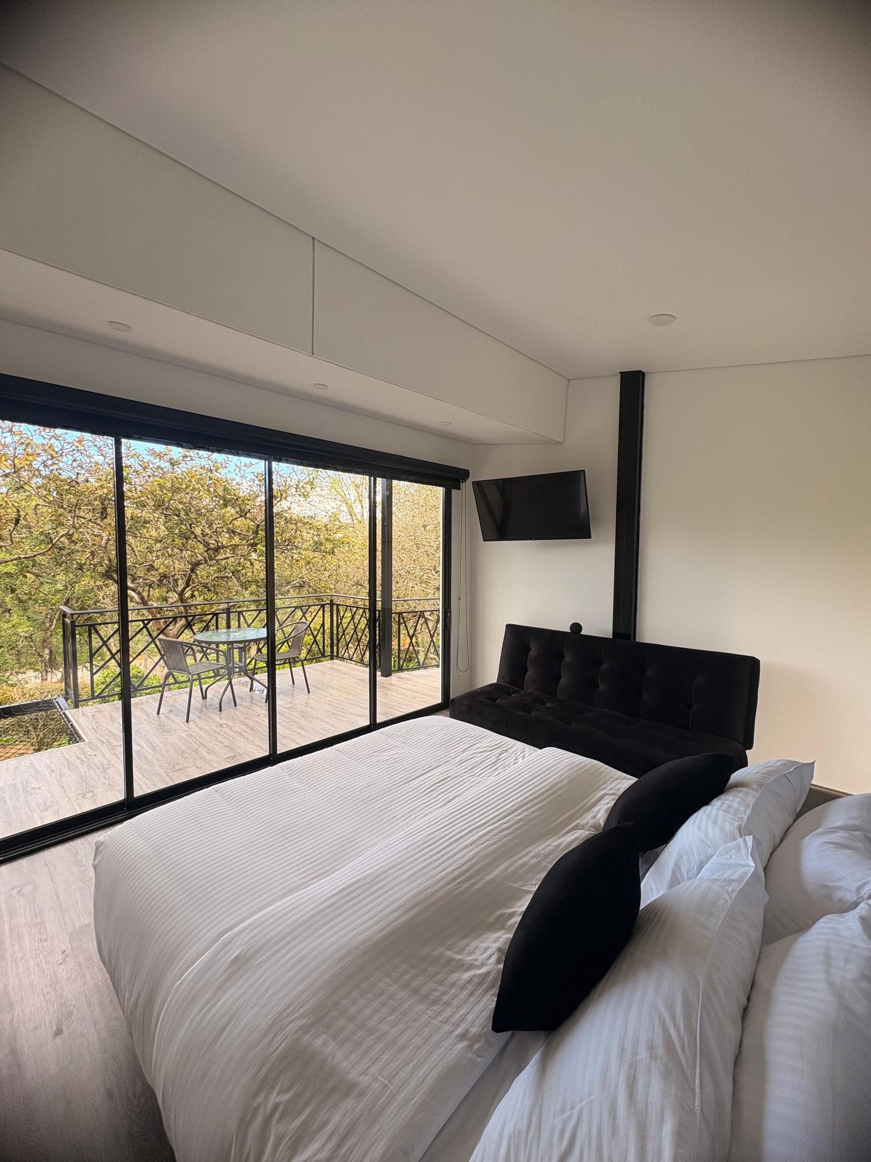 A modern bedroom with a large bed, a flat-screen TV mounted on the wall, and a balcony with a table and chairs.