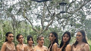 A group of women in formal attire standing on a wooden deck with a backdrop of trees.