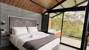 A modern bedroom with a large bed, a wooden ceiling, and a view of greenery outside.