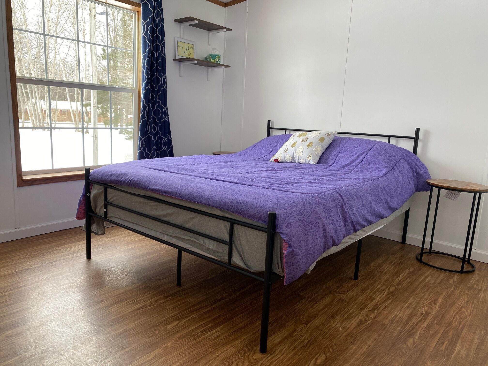 Een eenpersoonsbed met een paars dekbed in een kamer met houten vloeren, een raam met uitzicht op bomen, en een klein tafeltje naast het bed.
