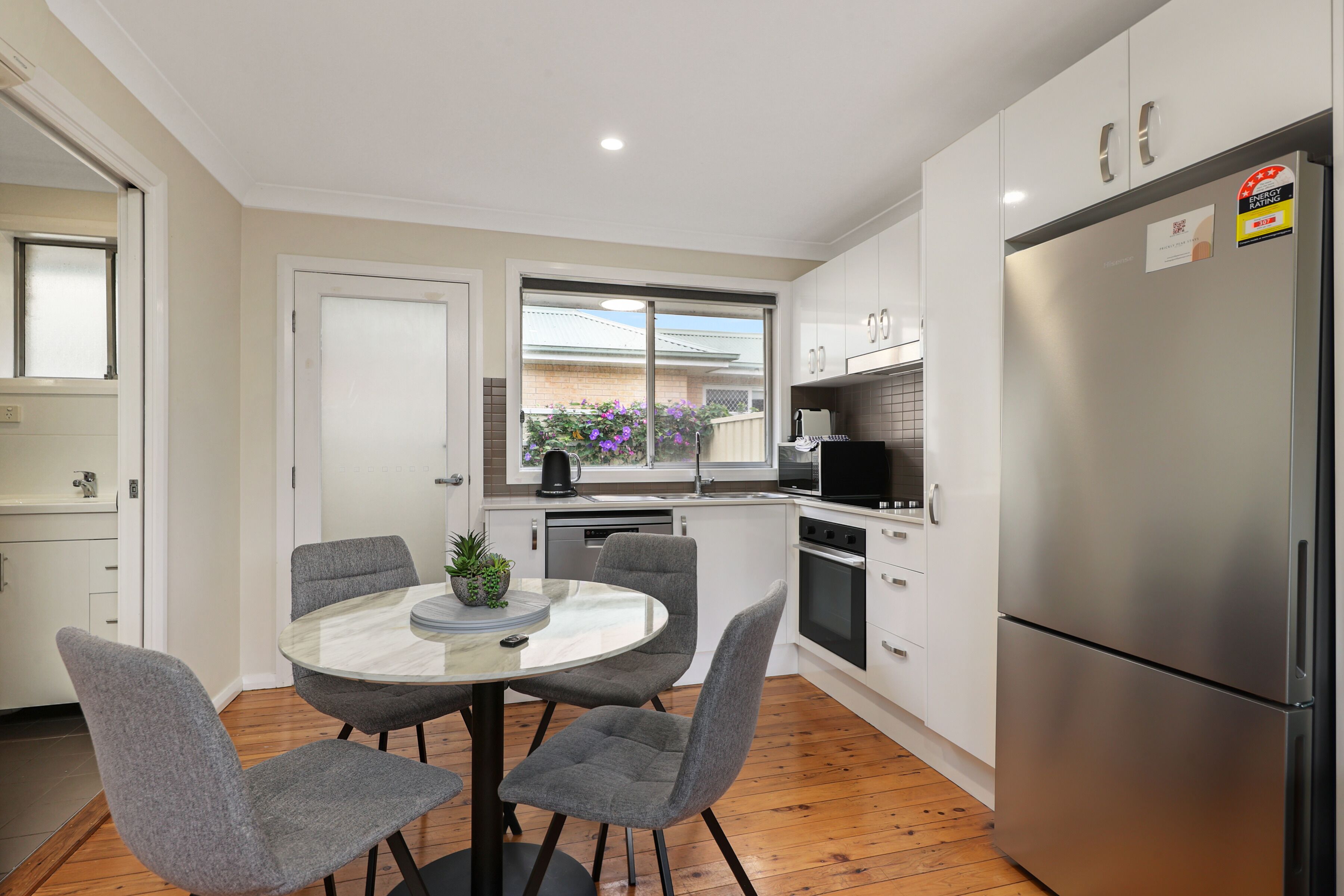 A modern kitchen with white cabinets, a stainless steel refrigerator, a dining table with four chairs, and a window with a view of a house and flowers outside.