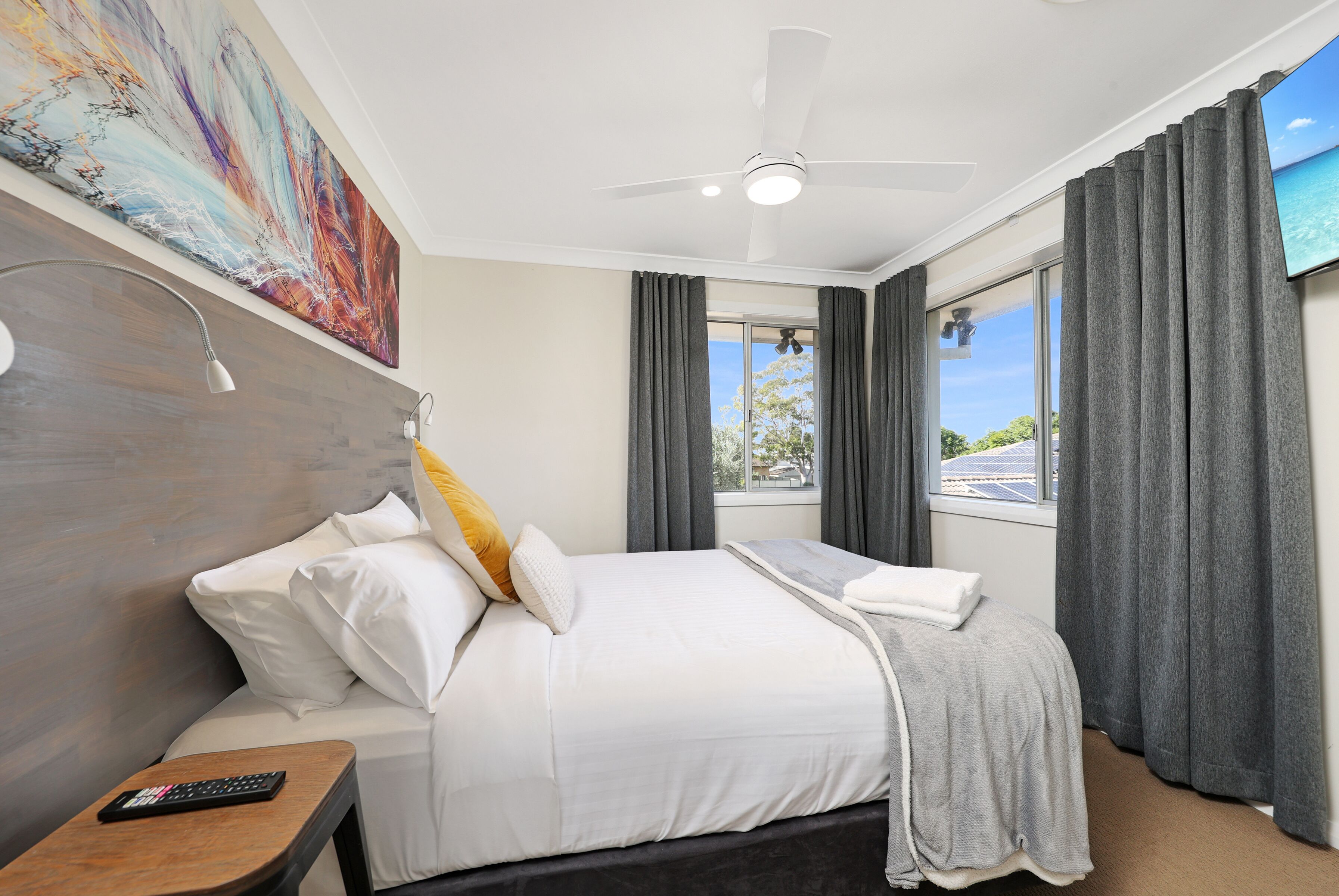 A hotel room with a bed, a nightstand, a ceiling fan, a large window with curtains, and a painting on the wall.
