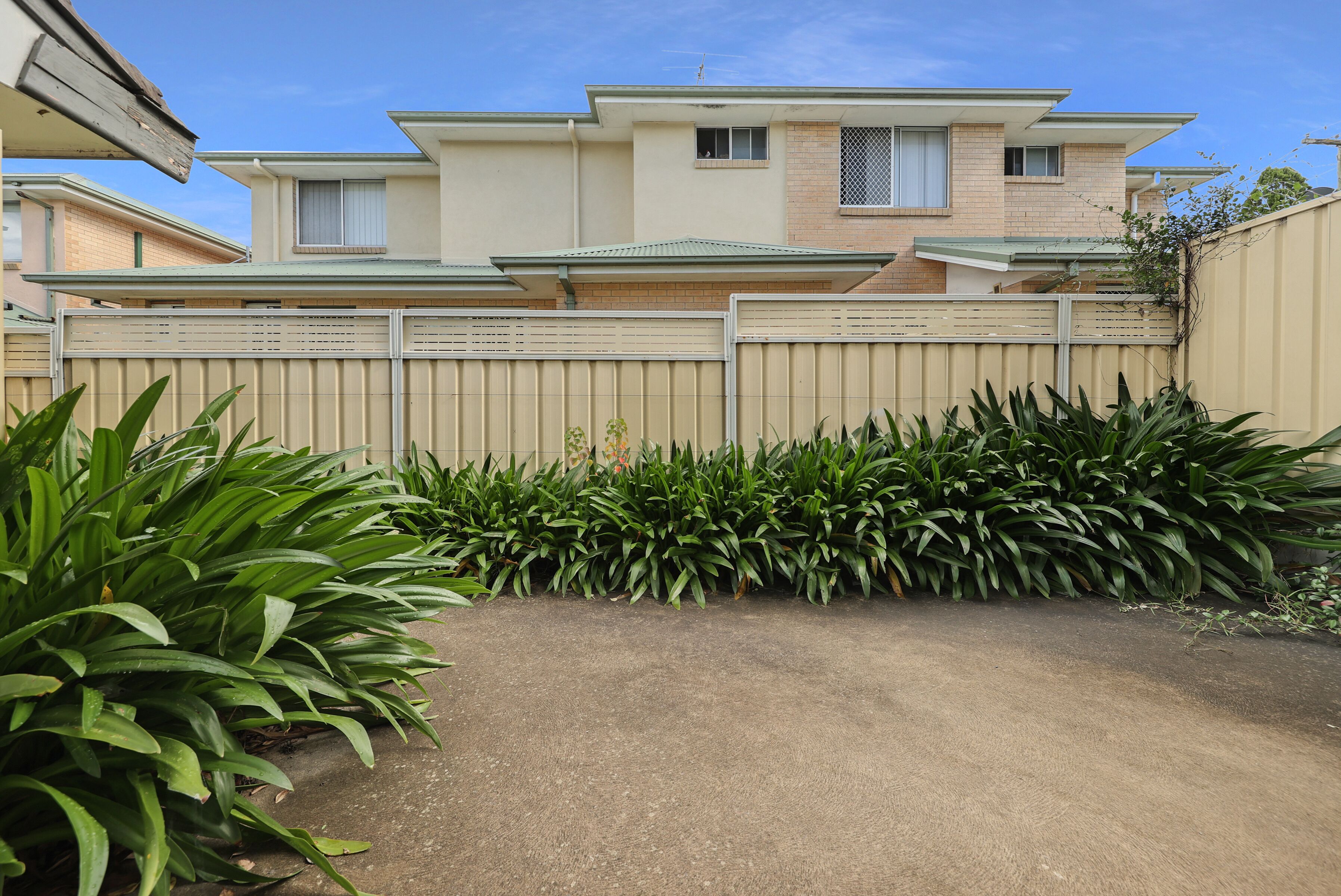 A residential house with a beige exterior, a gated driveway, and lush green plants in the foreground.