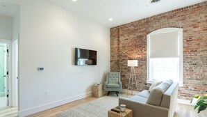 A modern living room with a brick wall, a flat-screen TV, a grey armchair, a sofa, and a floor lamp.