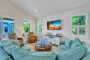 Amplia sala de estar con un sofá grande color turquesa, un sillón de cuero marrón, una mesa de centro de mimbre y un armario blanco con una pintura de atardecer tropical.