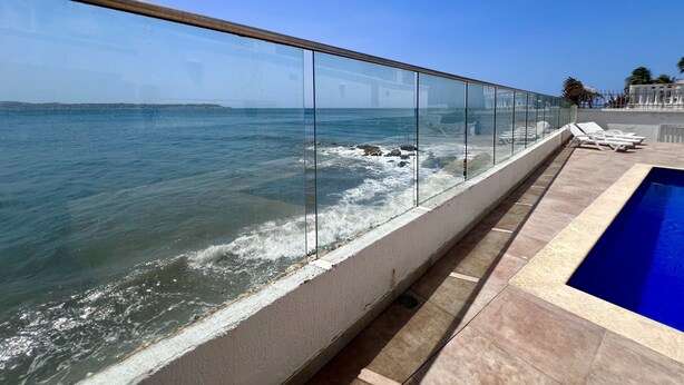 Área da piscina com guarda-corpo de vidro, vista para o mar, céu claro e costa distante.