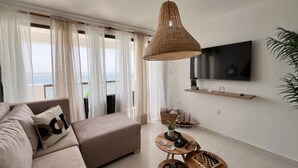 Uma sala de estar moderna com um sofá, uma TV fixada na parede, uma mesa de centro de madeira e vista para o mar através de grandes janelas.