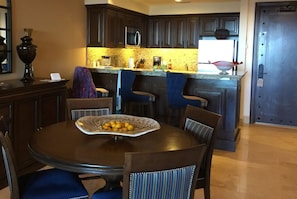 Un comedor con una mesa redonda, sillas y un frutero. Se observa una zona de cocina con armarios oscuros y un microondas.