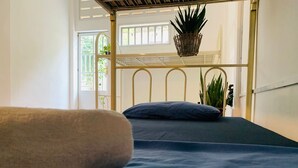 Un lit superposé avec un matelas bleu et une couverture beige, une tête de lit en métal et une fenêtre donnant sur un paysage verdoyant.
