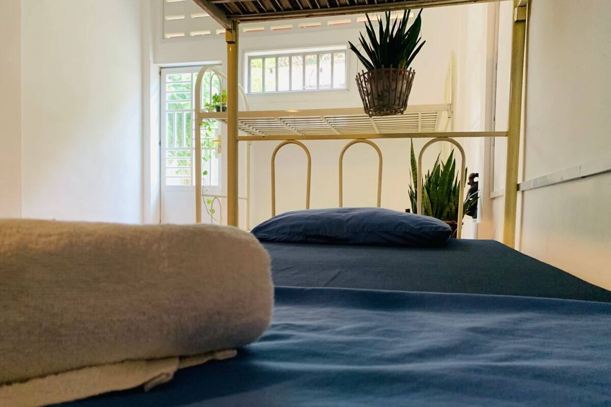 Een stapelbed met een blauwe matras en een beige deken, een metalen hoofdbord, en een raam met uitzicht op groen.