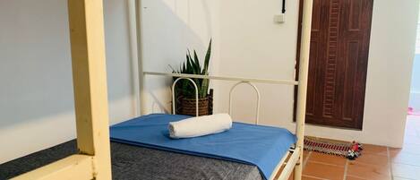 A single bed with a blue mattress and grey sheet, a white towel, and a potted plant on the nightstand.