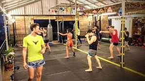 Une séance d’entraînement aux arts martiaux dans une salle de sport équipée de tapis, de sacs de frappe et d’un plancher en bois.
