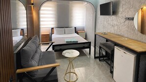 A compact hotel room with a bed, a sofa, a small table, a desk, a TV, and a mirror.