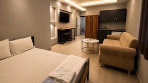 A modern hotel room with a bed, a sofa, a small round table, a TV, and a kitchenette.