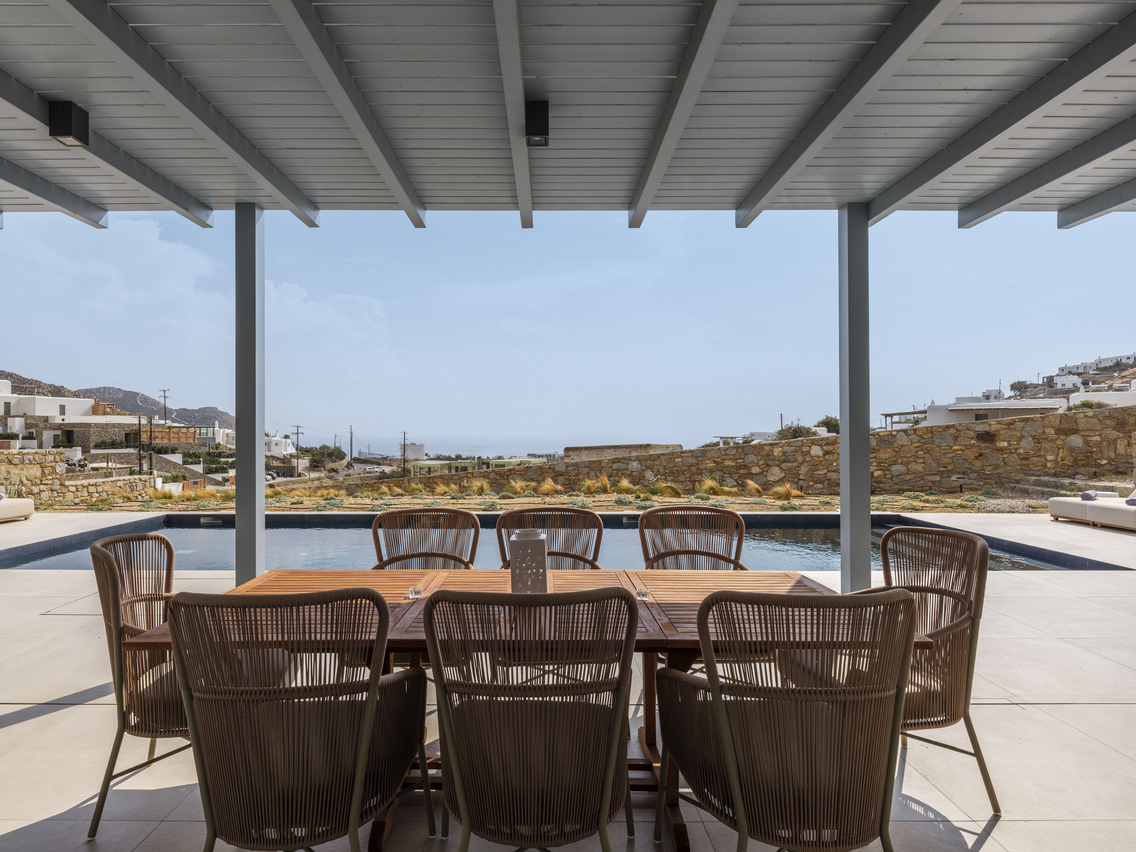 A covered outdoor dining area with a pool, wicker chairs, and a view of the coastline.