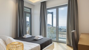 A modern hotel room with a large bed, a wooden side table, and a view of the sea through floor-to-ceiling windows.
