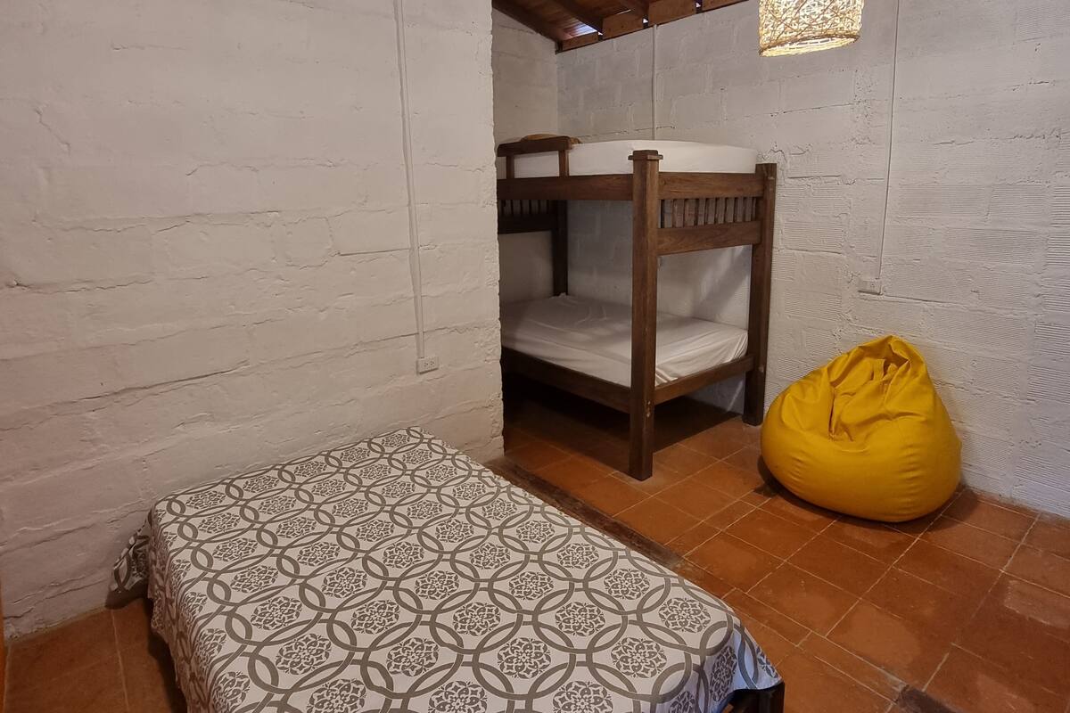 Una habitación con una cama litera de madera, una colcha estampada, un sillón de mimbre amarillo y dos lámparas colgantes.