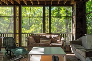A cozy room with a wooden ceiling, a sofa set, a table, and a view of a forest through large windows.