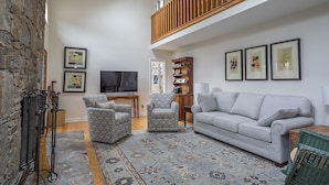 A spacious living room with a high ceiling, a stone wall, a wooden balcony, and a large window. - Heavenfell | Spacious Montreat Home | 1 Mile from Lake Susan & Conference Center! (Montreat)