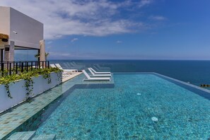 A clear blue infinity pool with white lounge chairs, a white building with a glass facade, and a view of the ocean. - Nomada Loft Zona Romantica Rooftop Infinity Pool (Puerto Vallarta)