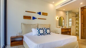Une chambre moderne avec un grand lit, une table de chevet en bois, un miroir et une salle de bain visible à travers une porte ouverte.