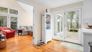 A spacious living room with a red armchair, a grey sofa, and a white cabinet with shelves. There are wooden floors and a large window providing a view of trees. - Sandy Spot: Walk to Beach, Sleeps 10 (Harwich Port)