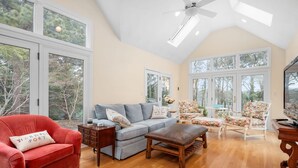 A spacious living room with a high ceiling, large windows, and a cozy seating area including a red armchair, grey sofa, and floral chairs. - Sandy Spot: Walk to Beach, Sleeps 10 (Harwich Port)