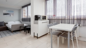 A modern hotel room with a bed, a sofa, a dining table with chairs, a kitchenette, and large windows with sheer curtains.