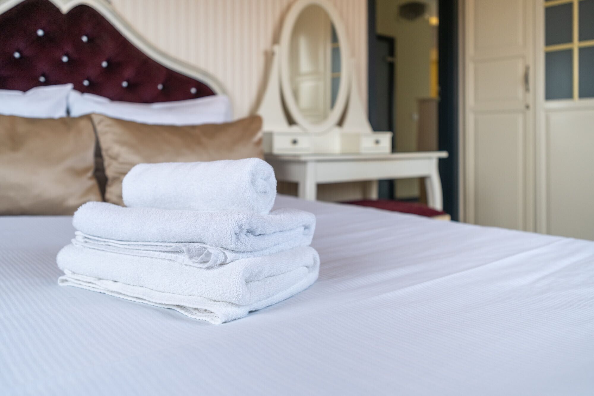 Una cama bien hecha con toallas blancas dobladas, un cabecero tapizado rojo y una mesita de noche blanca con espejo.