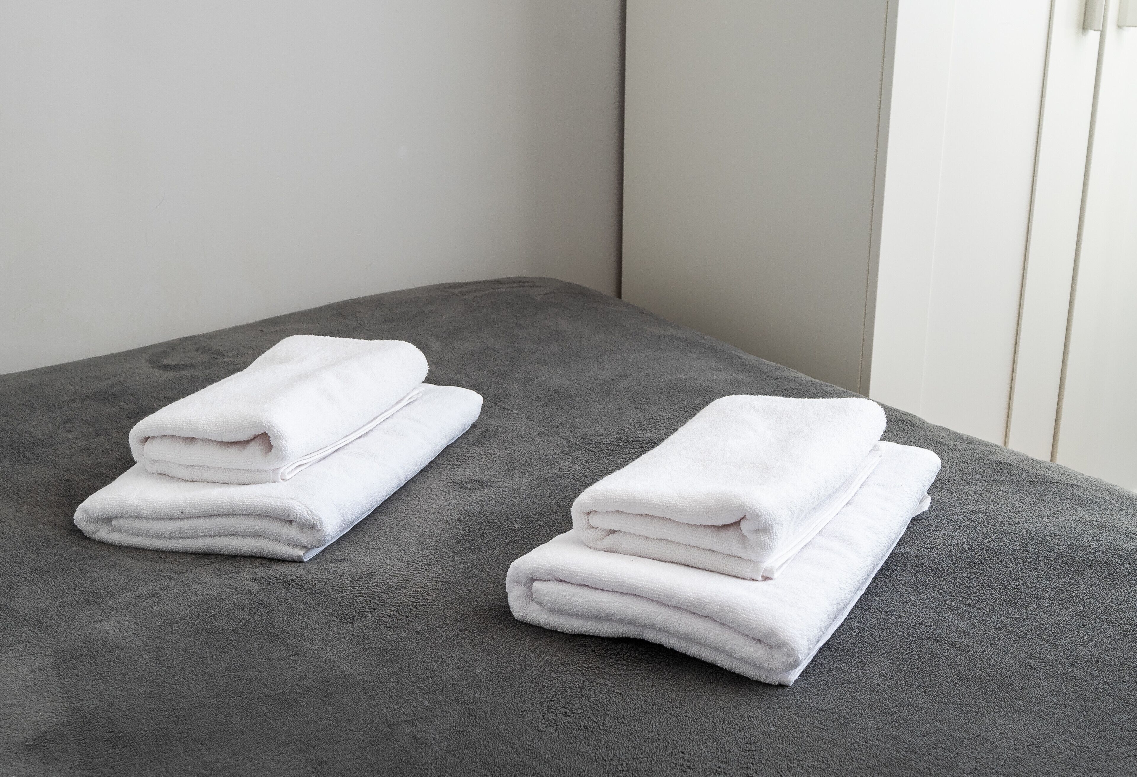 Una toalla blanca doblada cuidadosamente sobre una superficie gris con textura, probablemente un tapete de baño, en una habitación con una pared blanca lisa y un panel vertical.