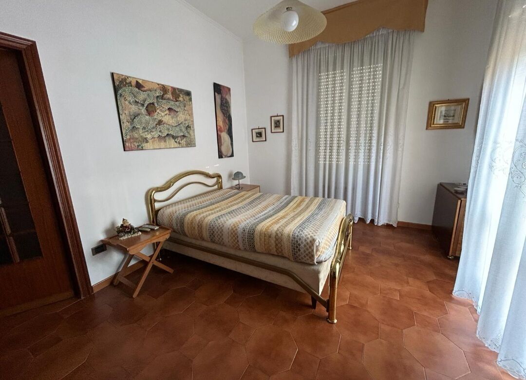 Ein Schlafzimmer mit Bett, Nachttisch, Lampe und einem Fenster mit Vorhängen.