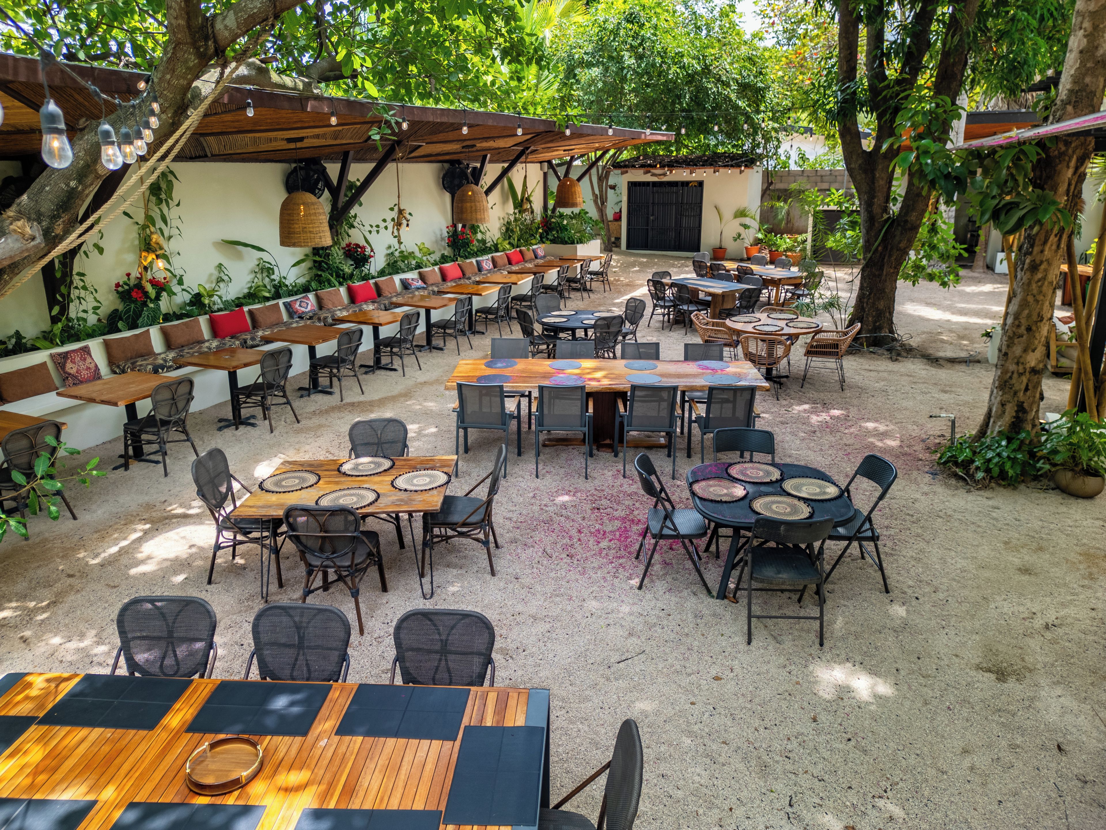 An outdoor dining area with multiple tables and chairs, surrounded by greenery and trees.