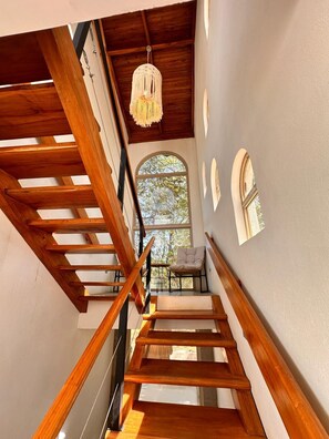 Una escalera de madera con un candelabro, un ventanal con vistas a árboles y una silla.