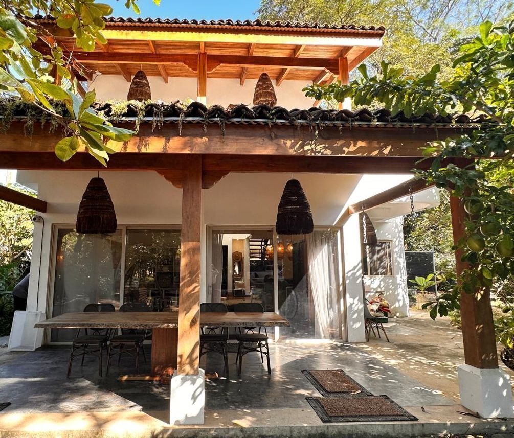 A covered outdoor dining area with a wooden table and chairs, a pergola overhead, and a view into the house.